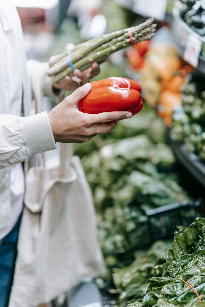 Crop anonymous buyer with shopping bag standing near stall with various greens with bell pepper and asparagus in hands during grocery shopping